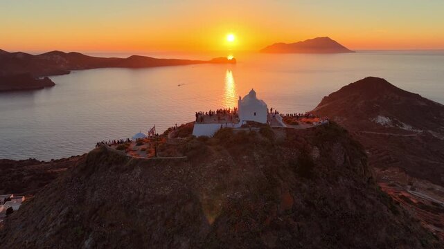 Idyllic drone seascape of Milos island in Aegean Sea, vacation in the Cyclades, aerial view of Greek traditional whitewashed church on a hill at sunset, drone is descending behind the church