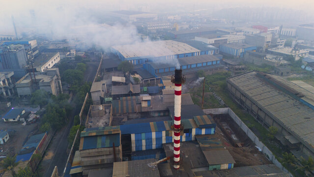 Aerial view of the Sachin industrial area, featuring factory buildings with rooftops and a tall red and white striped chimney emitting smoke into the hazy sky in Surat, Gujarat, India.