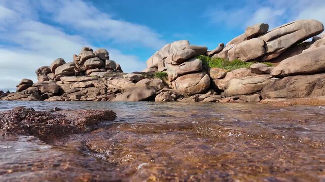Low-angle view of shallow seawater moving over coastal rocks toward massive rounded granite boulders beneath a blue sky in slow motion