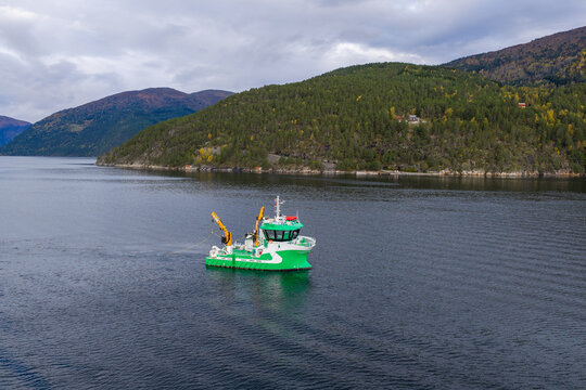 Aerial view of a green service vessel operating an underwater drone for inspection in a fjord, surrounded by forested mountains in Liabygda, More og Romsdal, Norway.