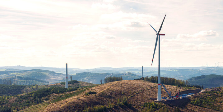 Aerial view of a wind turbine park under construction with tall white towers and heavy machinery on a hilly landscape in Germany.