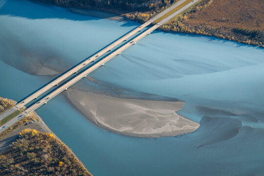 Aerial view of the Knik River bridge crossing glacial blue waters with a prominent sandbar and surrounding autumn foliage Knik River, Alaska, United States.