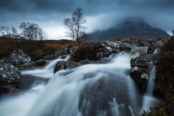 A lone birch stands beside a cascading waterfall, with Buachaille Etive Mòr disappearing into swirling cloud behind. This is one of Scotland's most iconic mountain views. © Scott