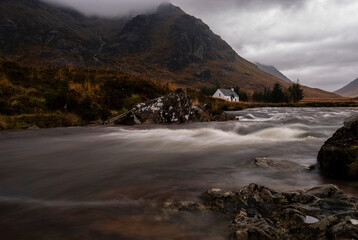 A fast-flowing river leads towards the iconic white cottage of Lagangarbh, sitting at the foot of Buachaille Etive Mòr under heavy, moody cloud. © Scott