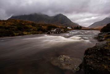 A fast-flowing river leads towards the iconic white cottage of Lagangarbh, sitting at the foot of Buachaille Etive Mòr under heavy, moody cloud. © Scott