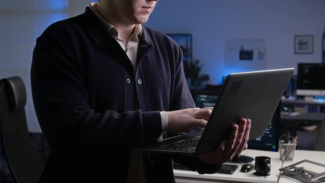 Caucasian young adult man holding laptop while typing code in modern office. Footage ideal for cybersecurity tutorials, IT development marketing and corporate data protection promos