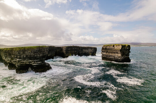 Aerial view of the dramatic sea cliffs at Downpatrick Head with crashing waves, layered rock formations, and green grassy tops under a cloudy sky in Lackan South, Mayo, Ireland.