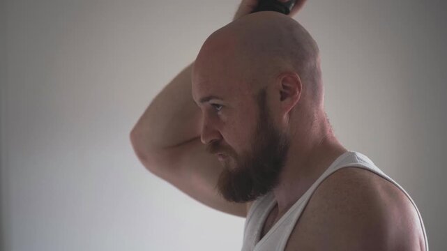 Barbershop at home and razor ad, bald man with beard in white tank top shaving back of head on grey background. Barbershop service concept for men, man turns head and shaves nape with trimmer