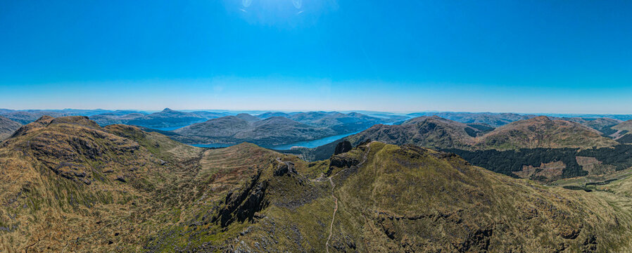 Aerial view of the rugged mountainous landscape of the Scottish Highlands with rolling green hills, rocky peaks, and a distant lake in the United Kingdom.