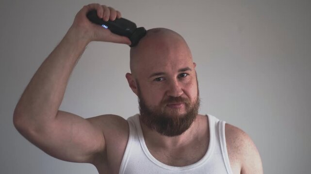 Hair loss and alopecia concept, bald bearded man in white tank top shaving head with clipper on grey background. Alopecia acceptance and men health ads, man shaves crown and looks at camera