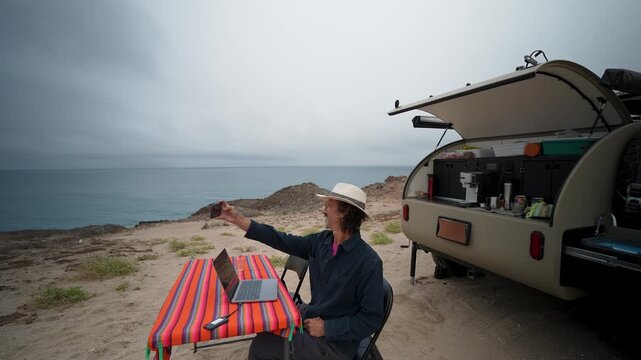 A senior man works on his laptop and takes selfies outdoors by his teardrop camper while boondocking in beautiful Baja Mexico.