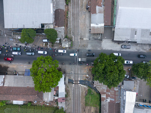 Aerial view of a railway crossing with cars and motorcycles waiting on the road amidst urban buildings and lush trees in Indonesia.
