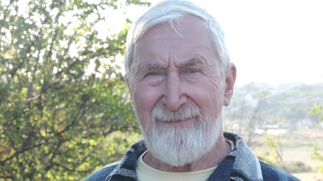 Dental care and denture ad, smiling senior man with white beard in denim shirt outdoors among trees. Healthy smile in old age concept, elderly man smiles at camera
