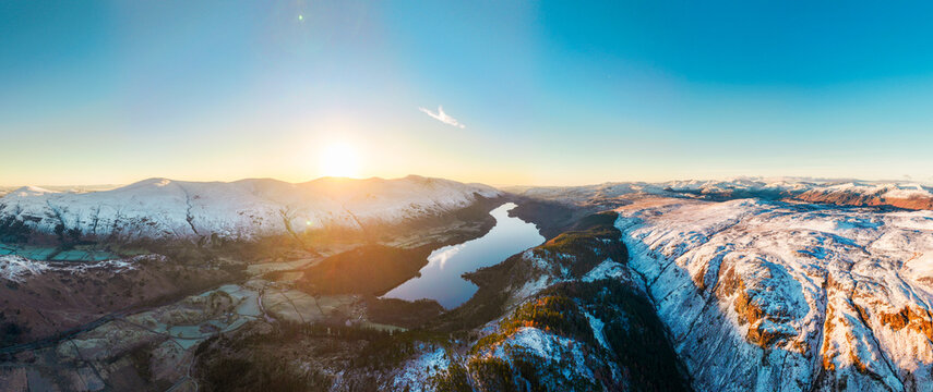 Aerial view of Lake District National Park featuring snow-covered mountain peaks, a calm lake reflecting the golden sunrise, and rugged terrain in England, United Kingdom.