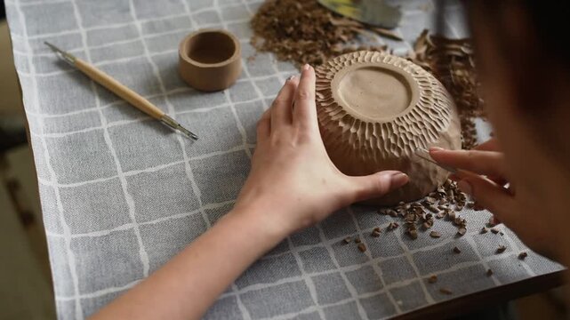 Textured ceramic bowl making, hands carving pattern on clay bowl with wooden tool on checkered table. Handmade tableware concept, potter carves texture on bowl