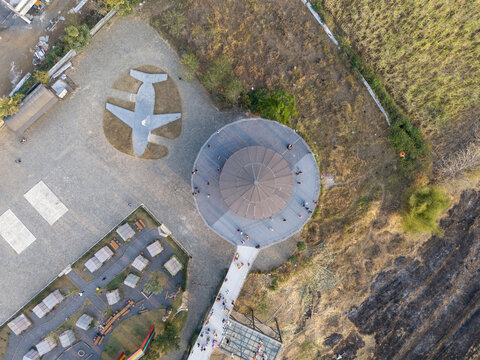 Aerial view of Malang Skyland with its circular building, airplane-shaped ground marking, and glass bridge, surrounded by rural landscape in Kecamatan Karang Ploso, Jawa Timur, Indonesia.