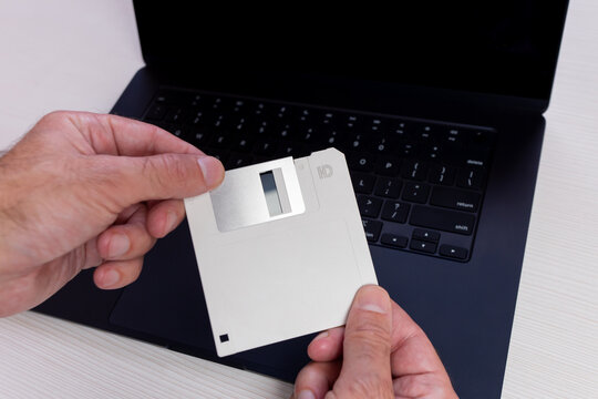 A person is holding a 3.5-inch floppy disk in front of a modern laptop.