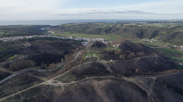 Aerial view of the village of Odeceixe surrounded by rolling hills with burnt vegetation and winding roads leading towards the Atlantic coast in Odeceixe, Faro District, Portugal.