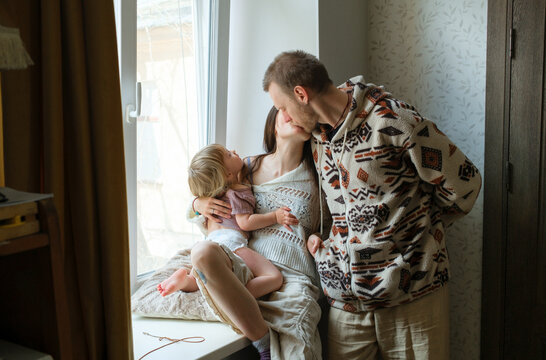 Parents kissing while sitting with their toddler on a windowsill