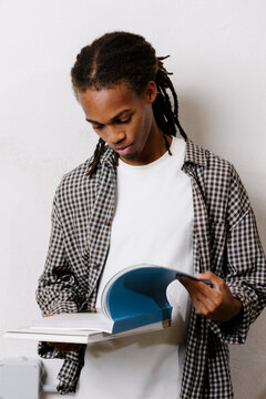 Young Black man with dreadlocks reading a book, white background.