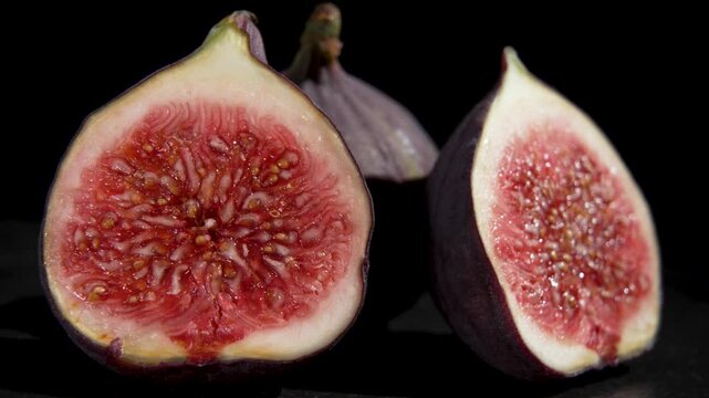 Two fig halves showing red seeded flesh slowly rotate against a black background, with a whole fig visible behind them.