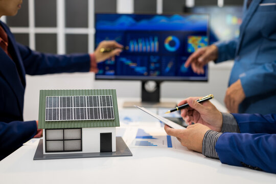 Business team reviewing solar energy plans with a miniature house, rooftop solar panels, charts, tablet and data dashboard in a modern office.