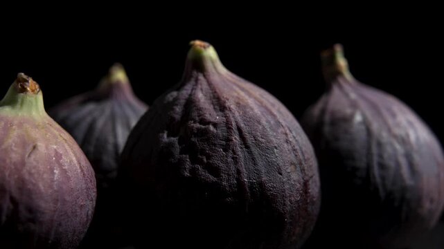Several whole ripe purple figs slowly rotate in low-key light against a black background, showing their stems and skin texture.