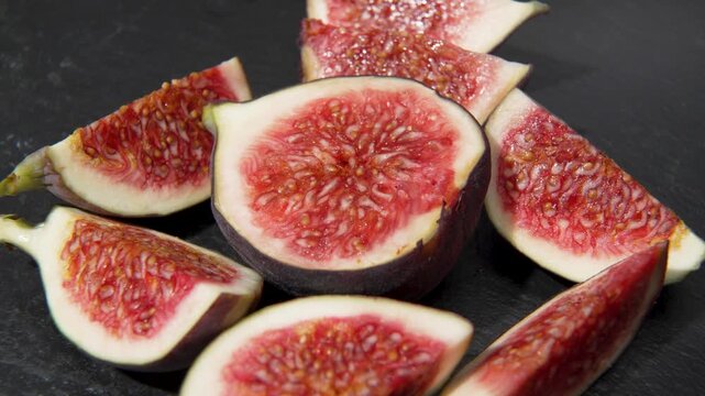 Fresh fig wedges arranged around a fig half slowly rotate on a dark slate surface, showing juicy red flesh and seeds.