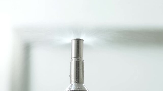 Metallic cylindrical nozzle releasing a fine water spray against a pale studio background