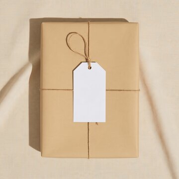 Blank gift tag on a kraft paper parcel with jute twine, featuring a fibrous texture on beige linen for a stationery mockup and copy space