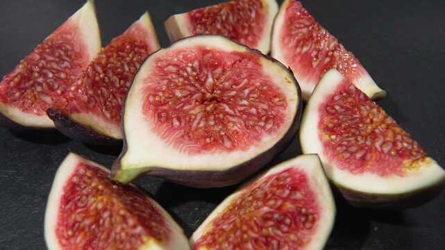 Top view of fresh fig wedges arranged around a fig half, slowly rotating on a dark slate surface. Red seeded flesh is visible.