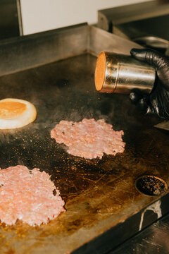 Chef seasons ground beef patties on a hot griddle in a commercial kitchen