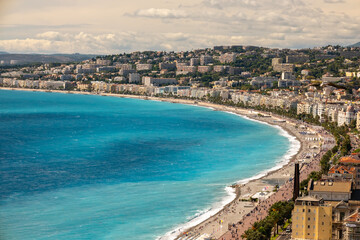 Fototapeta premium Mediterranean sea with turquoise water meeting the pebble beach and city buildings of nice, france, featuring the iconic promenade des anglais full of people