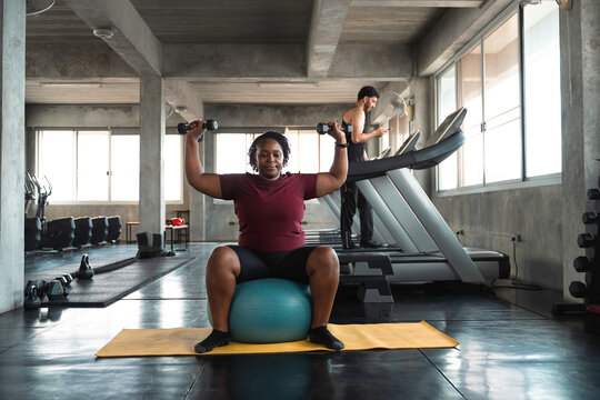 Focused curvy African American woman lifting dumbbells overhead while sitting on a blue stability ball in a modern industrial gym, demonstrating strength, health, and inclusive fitness motivation.