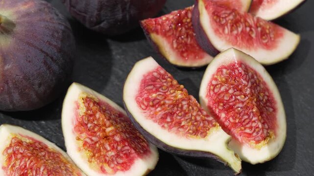 Close-up of fig wedges and whole purple figs slowly rotating on dark slate, showing juicy red flesh, seeds and stems.