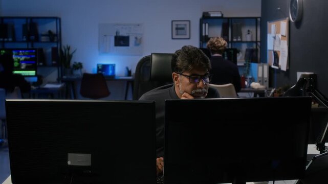 Middle aged South Asian cybersecurity analyst working on dual monitors in IT office. Perfect for corporate data protection, programming, and software development videos