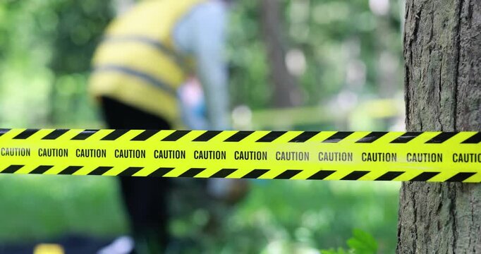 Yellow caution tape blocks off a wooded area while a blurred worker in a safety vest stands in the background.