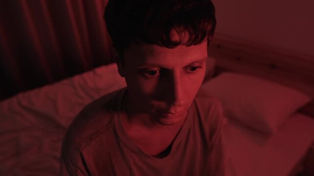 Anxious millennial man staring into the distance, lost in thoughts about life and uncertainty, portraying existential anxiety and emotional isolation in a dark red low key bedroom