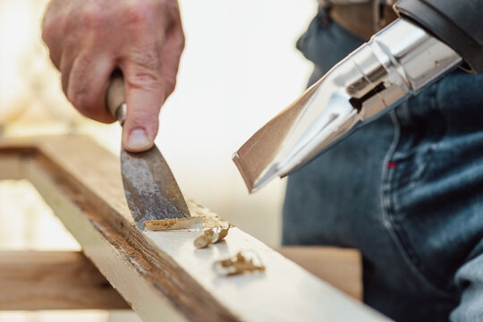 Carpenter using heat gun and wood scraper removes paint from old wooden window. Construction industry, carpentry. Restoration. 