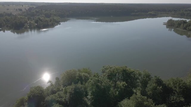 High aerial drone video panning over quiet lake with sun glare reflection and forest island near Lychen and Himmelpfort, Uckermark region, Brandenburg, Germany under clear blue sky on sunny day