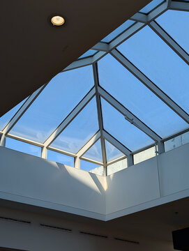 Geometric skylight structure providing natural light in a modern building interior, with a recessed ceiling spotlight and white walls.