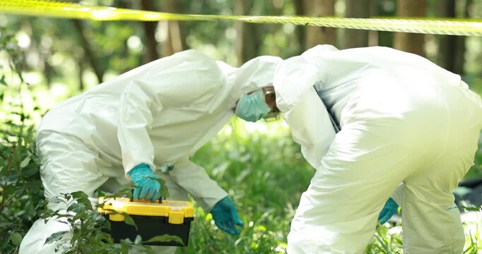 Two forensic investigators in protective suits, masks and gloves examine evidence in a wooded area behind yellow caution tape, with a toolbox nearby.