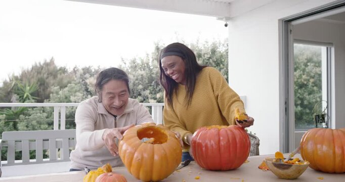 Asian senior man African American woman hugging then gutting and carving pumpkins on porch for fall