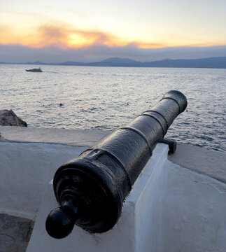 Black iron cannon overlooking rippled sea and mountain silhouettes at sunset in Hydra, Greece
