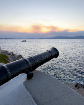 Black iron cannon overlooking coastal sea and motorboat at sunset in Hydra, Greece