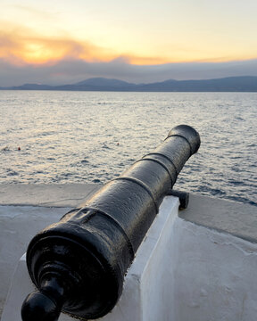 Black iron cannon overlooking rippling water and distant mountains at sunset in Hydra, Greece