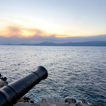 Black iron cannon overlooking rocky waterfront and distant hills at sunset in Hydra, Greece