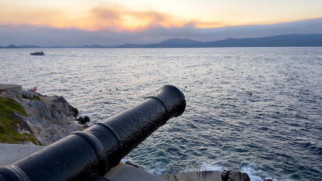 Weathered iron cannon points over rocky coast at sunset in Hydra, Greece