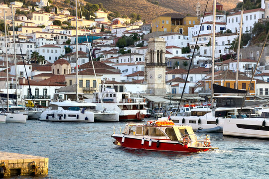 Red motorboat passes moored yachts beneath stone clock tower and hillside homes in Hydra, Greece