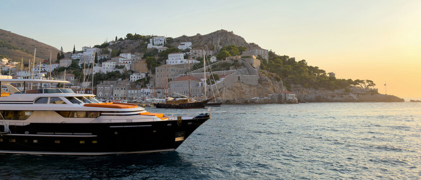 Luxury motor yacht in Hydra harbor, Greece, beside sailboats and hillside buildings at sunset
